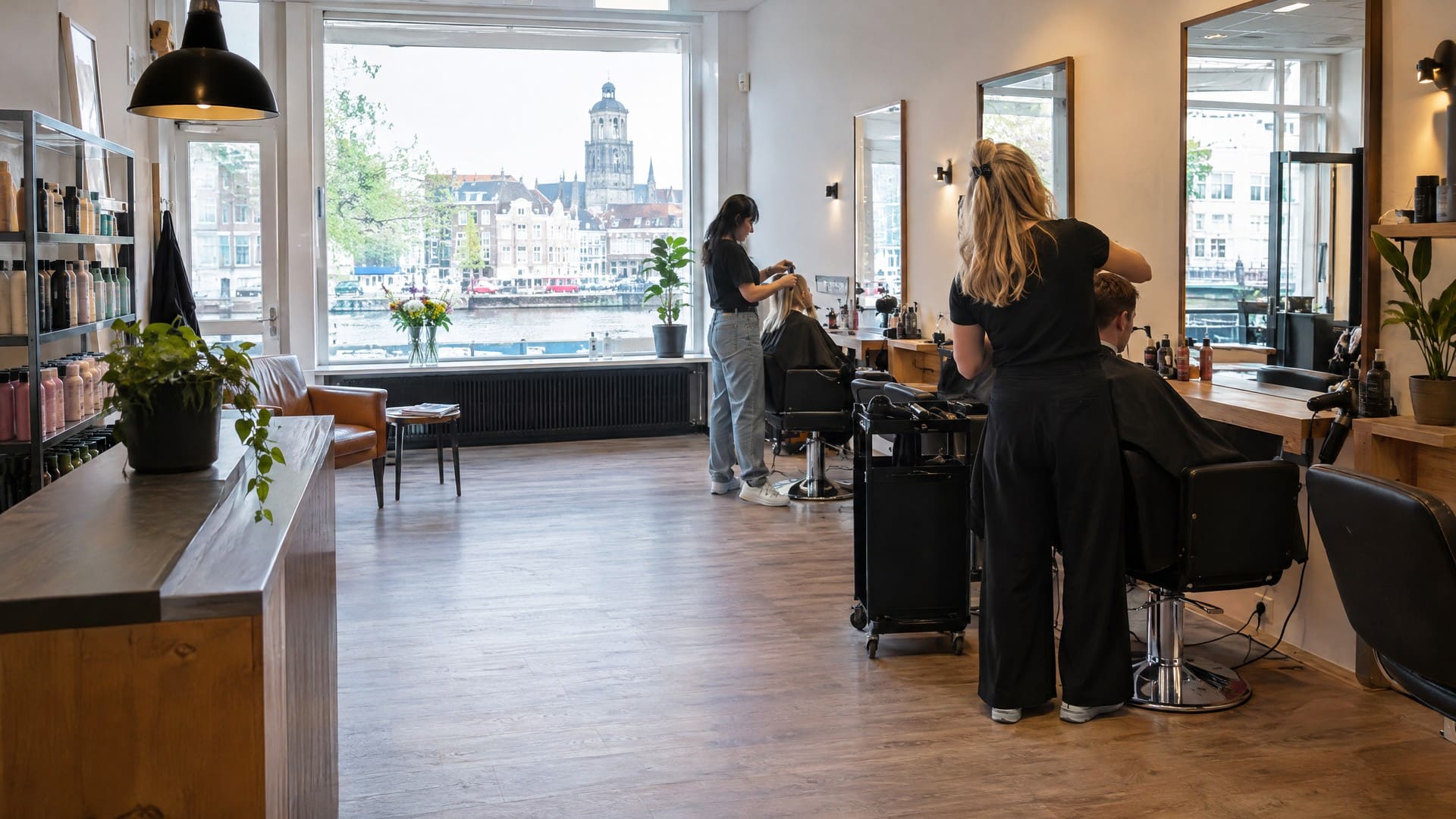 Ruime, lichte kapsalon met kapstoelen bij een groot raam met uitzicht over het water op de stad.
